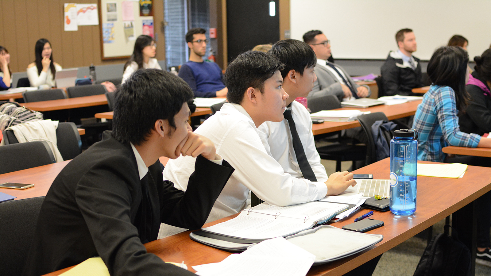 Students sit at desks listening attentively during a class, with notebooks, laptops, and papers on the tables