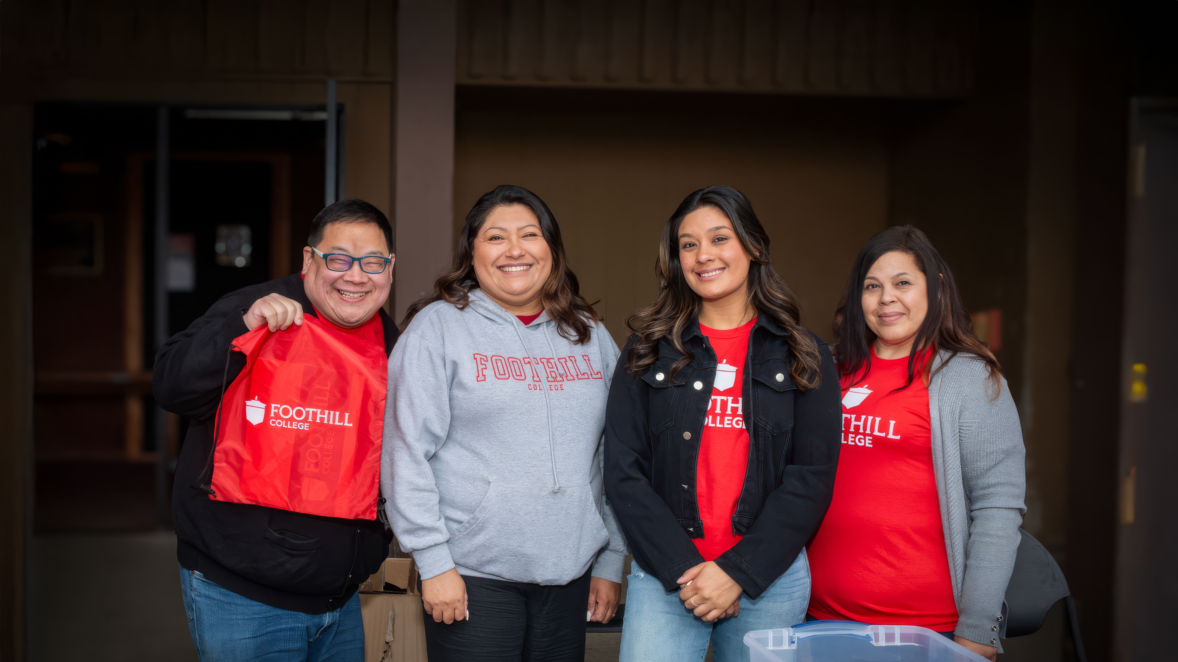 Four Foothill College staff members smiling and standing behind an information table, wearing red Foothill College shirts and hoodies, with outreach materials displayed in front of them.