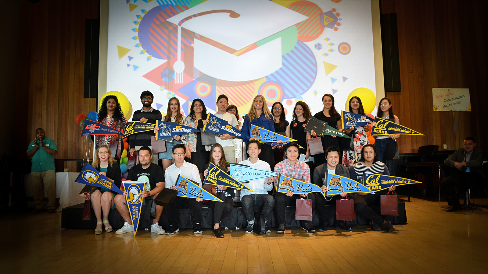 Group of transfer students holding university pennants on stage at a graduation celebration.