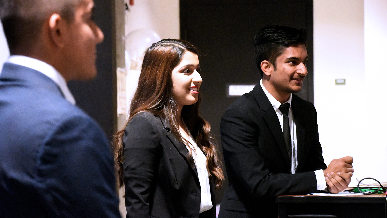 Three young adults dressed in formal business attire smile and talk together indoors during a professional event