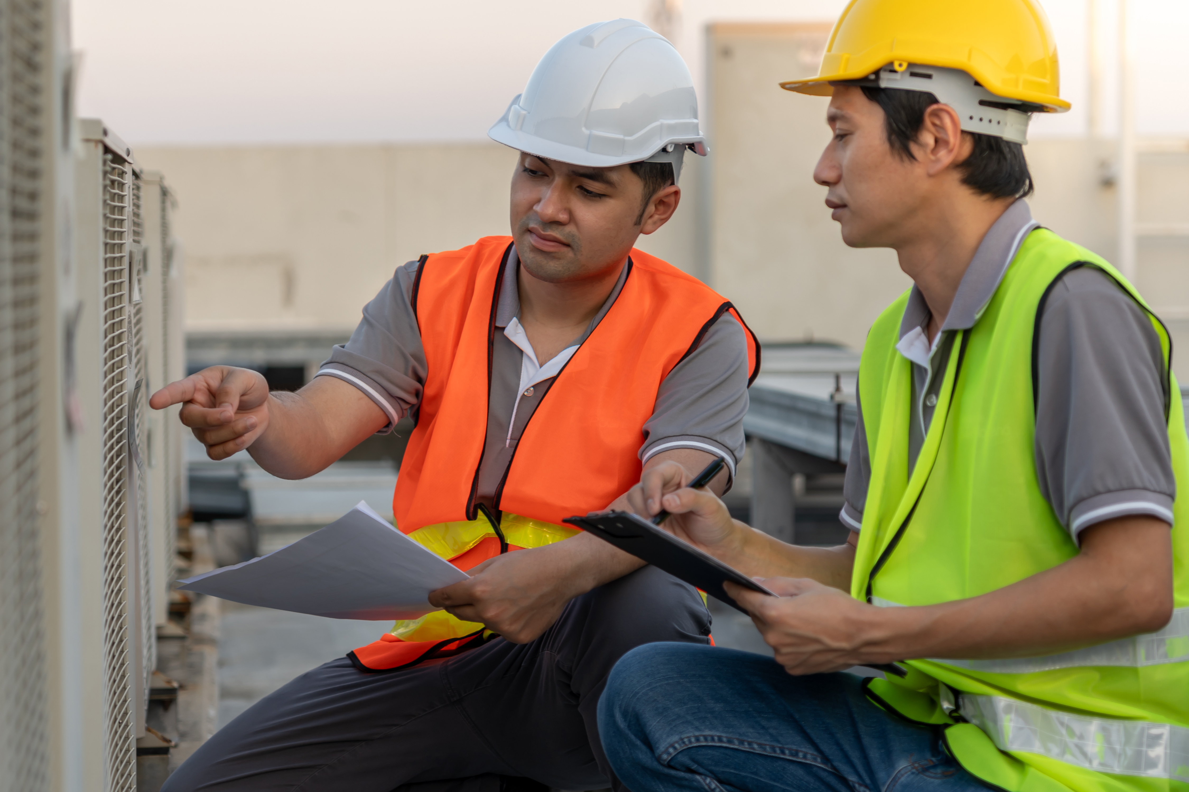 Two workers in safety vests and hard hats review HVAC equipment and notes.