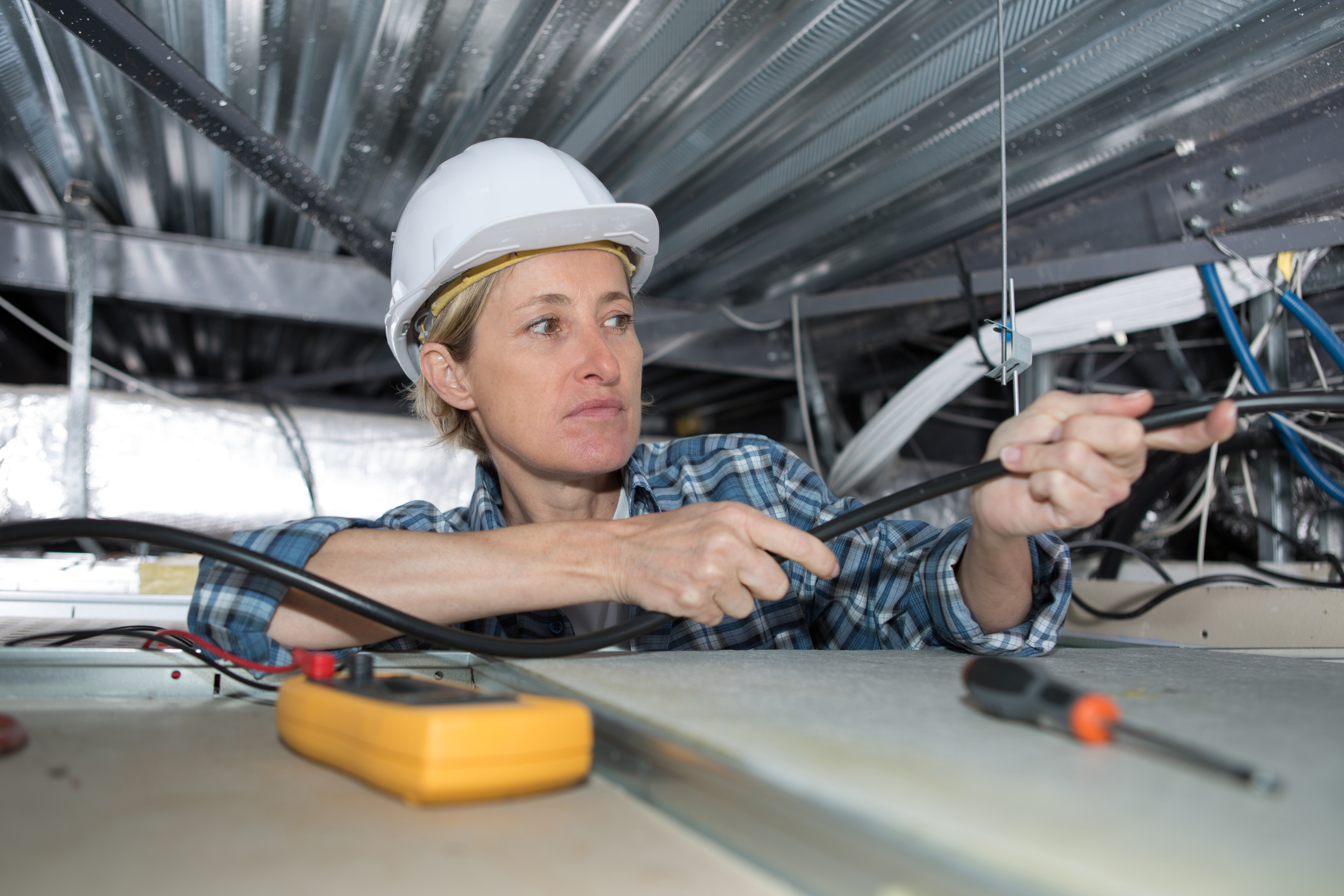 Electrician in a hard hat works in a ceiling space, pulling electrical cable.
