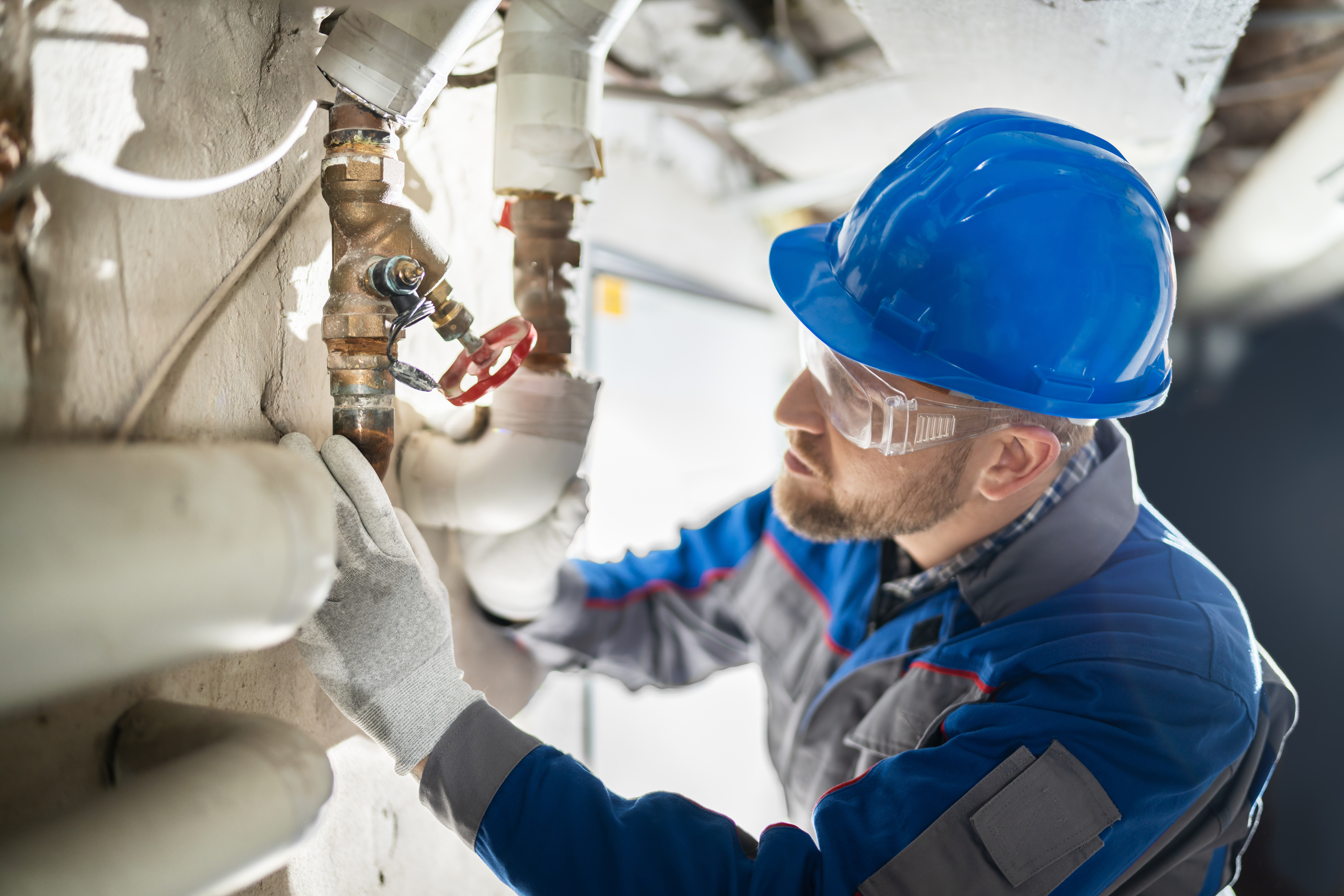 Worker in protective gear inspects plumbing pipes and a valve.