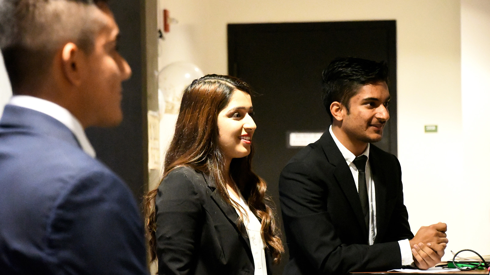 Three young adults dressed in formal business attire smile and talk together indoors during a professional event