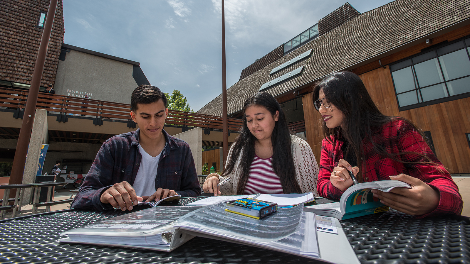Three students study together at an outdoor table on the Foothill College campus, reading books and taking notes in natural daylight.