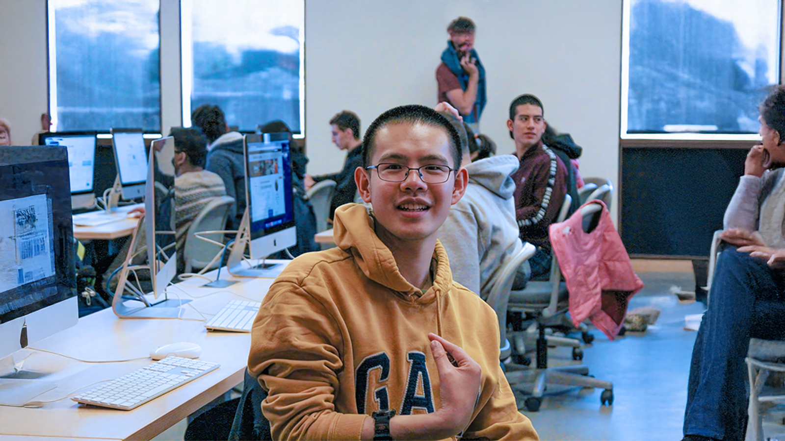 Student smiling in a computer lab classroom with other students working at desktop computers