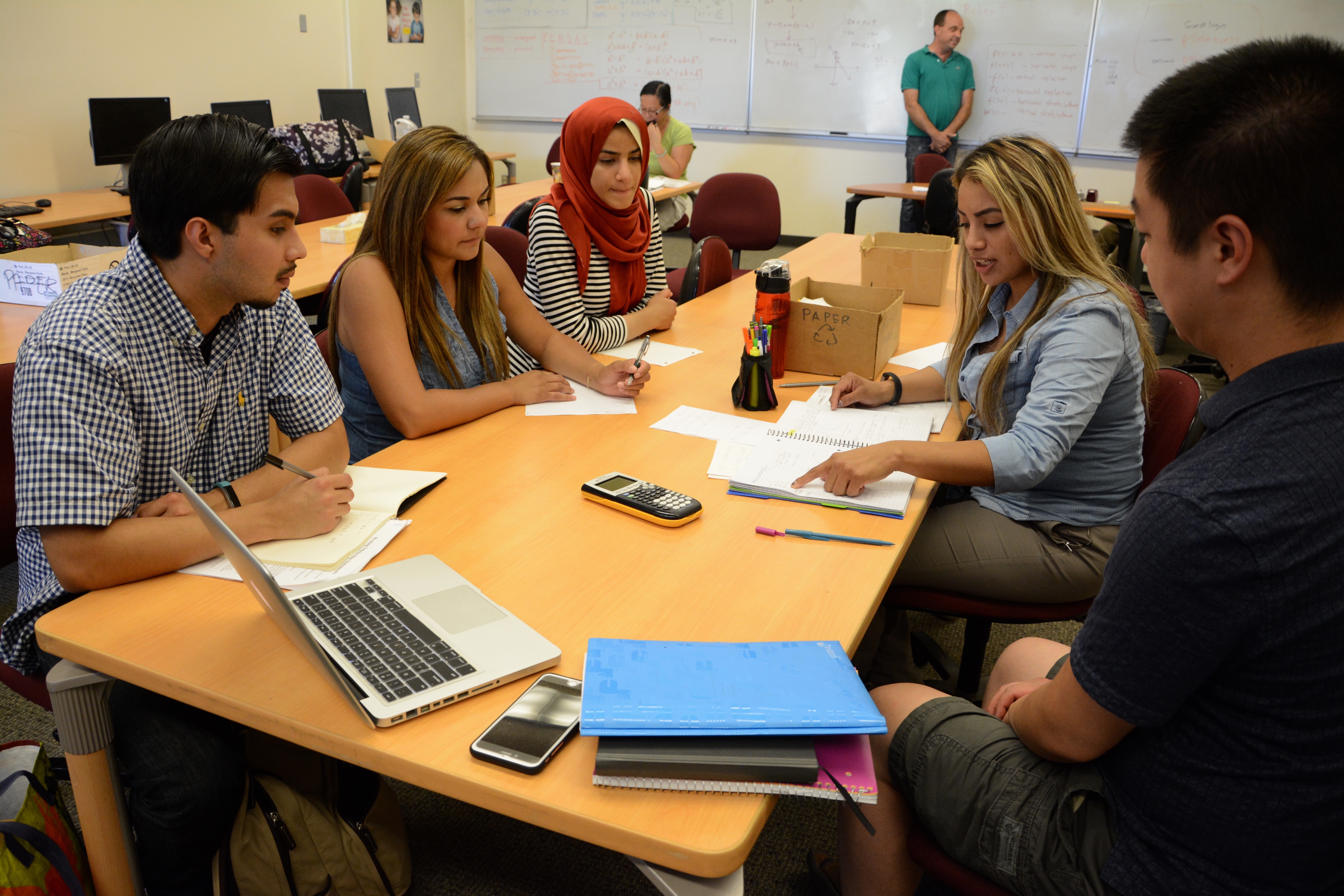 Students sit around a table in a classroom working together on a group assignment with laptops, notebooks, and calculators.
