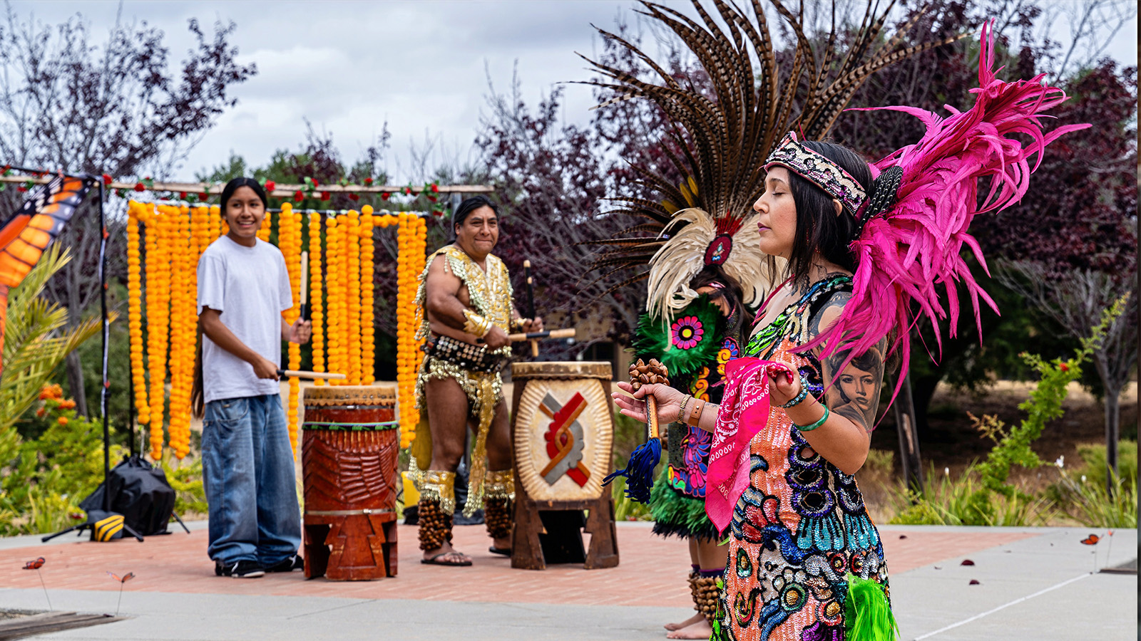 Cultural dance performance outdoors with performers in traditional attire, featuring drumming, vibrant costumes, and decorative marigold displays.