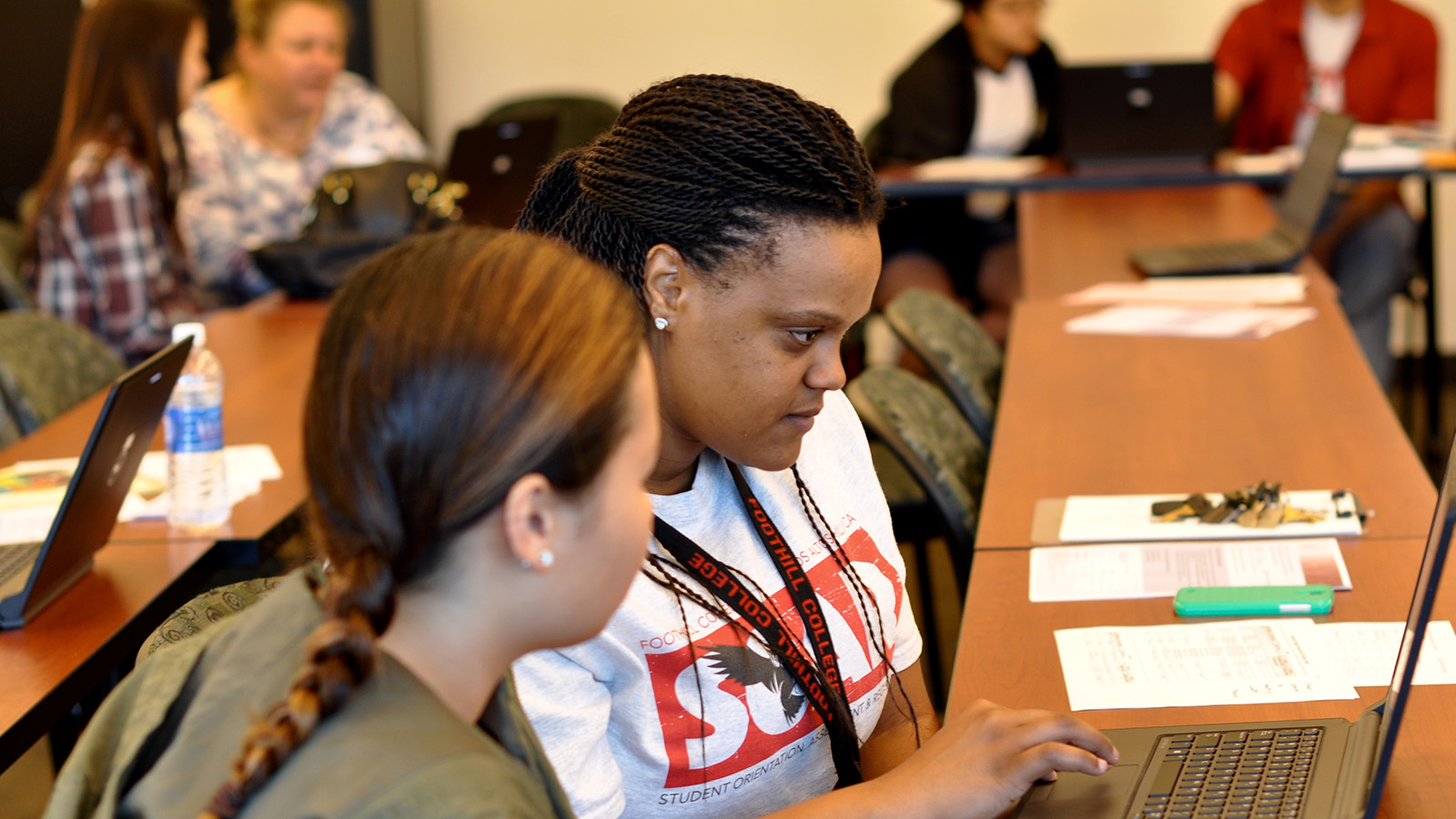 Two students sit together in a classroom, working on a laptop and reviewing papers.