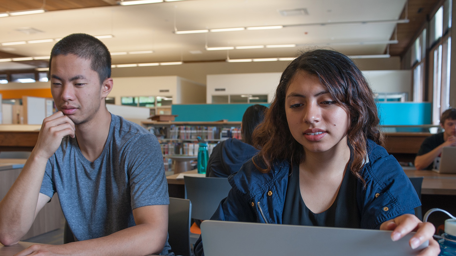 Two students work together on a laptop in a library study area.