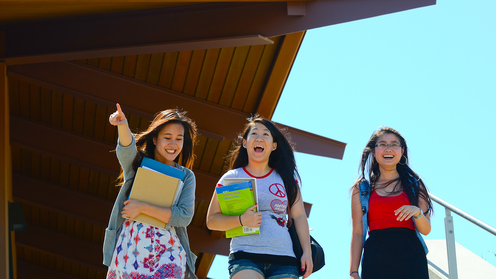 Three students walk on campus smiling and talking, carrying books and backpacks.